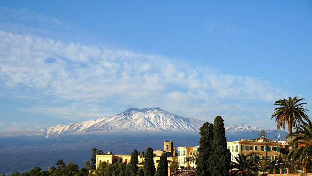 Snow-capped mount etna overlooking a sicilian town with lush greenery.